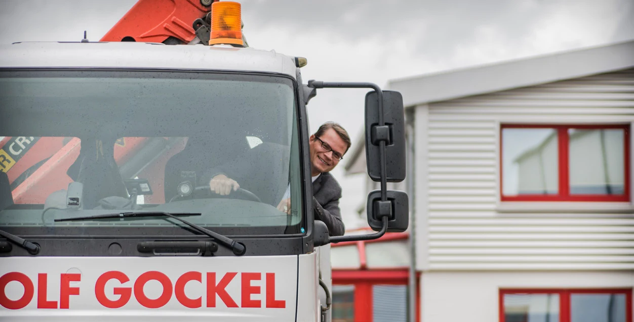 LKW mit Ladekran und Schriftzug Meinolf Gockel vor dem Firmengebäude, der Fahrer schaut aus dem Fenster